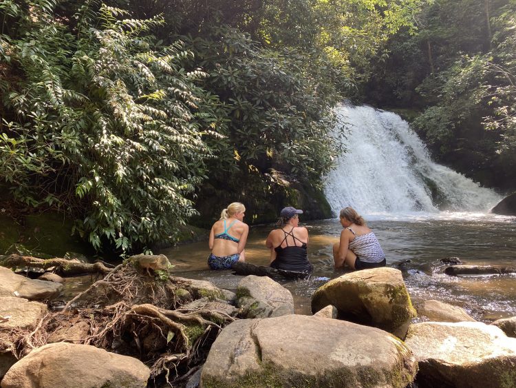 Three people sitting at the edge of a pool at the base of a waterfall.