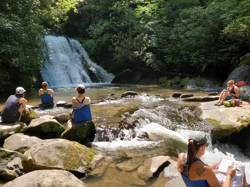 Five people sitting scattered on rocks around a pool at the base of a waterfall