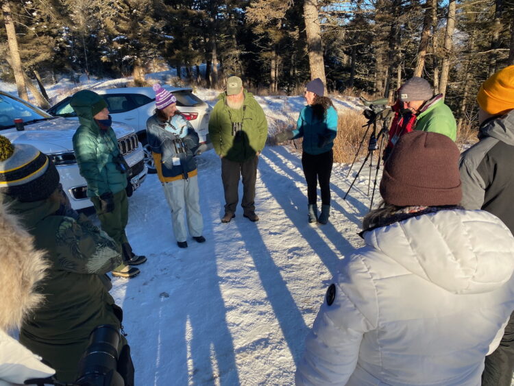 A group of teachers stands outdoors in a circle with two biologists that study wolves. 